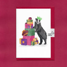 Load image into Gallery viewer, A hand drawn illustration on a greeting card of a festive badger wearing a paper Christmas hat and standing besides a pile of Christmas presents. The card is on top of festive red envelope and held together with a 100% plastic free card catch sticker
