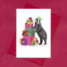 Load image into Gallery viewer, A hand drawn illustration on a greeting card of a festive badger wearing a paper Christmas hat and standing besides a pile of colourful Christmas presents. The card is on top of festive red envelope, sitting on a red background
