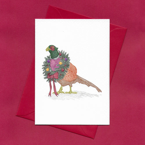 A hand drawn illustration on a greeting card of a festive pheasant wearing a colourful Christmas wreath around its neck. The card is on top of festive red envelope, sitting on a red background