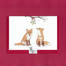 Load image into Gallery viewer, A hand drawn illustration on a greeting card of two foxes sitting beneath a bunch of mistletoe tied with a red bow and hanging from a branch. The card is on top of festive red envelope and held together with a 100% plastic free card catch sticker, sitting on a red background 
