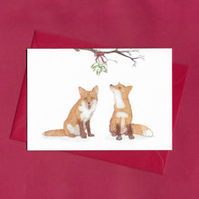 Load image into Gallery viewer, A hand drawn illustration on a greeting card of two foxes sitting beneath a bunch of mistletoe tied with a red bow and hanging from a branch. The card is on top of festive red envelope, sitting on a red background 
