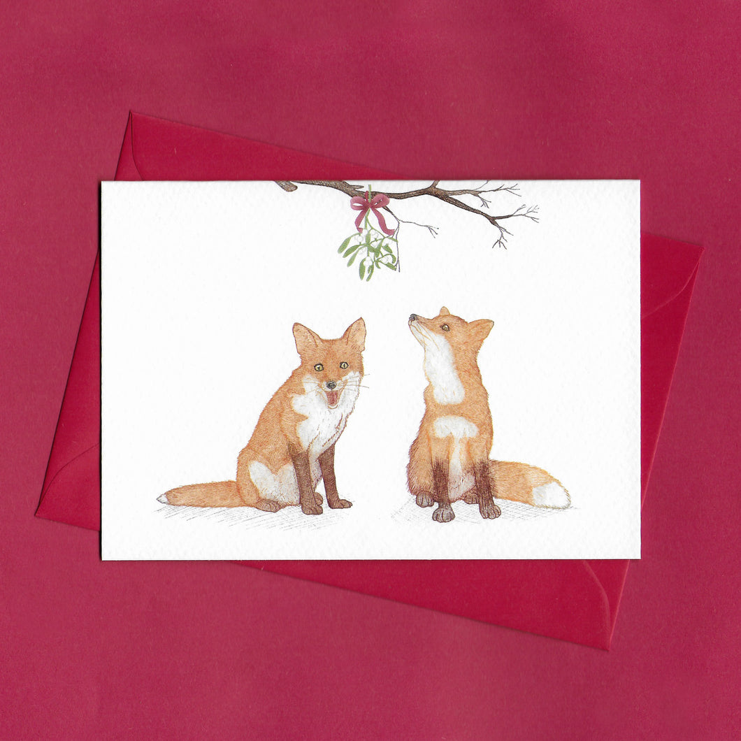 A hand drawn illustration on a greeting card of two foxes sitting beneath a bunch of mistletoe tied with a red bow and hanging from a branch. The card is on top of festive red envelope, sitting on a red background 