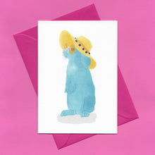 Load image into Gallery viewer, A stylised watercolour illustration of a blue rabbit wearing sunglasses and a yellow floppy sunhat on a greeting card, with a bright pink envelope, on a pink background.
