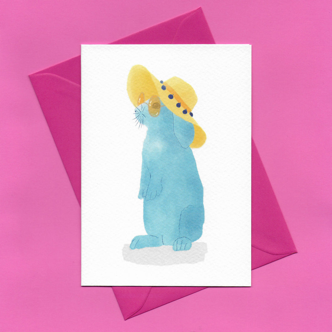 A stylised watercolour illustration of a blue rabbit wearing sunglasses and a yellow floppy sunhat on a greeting card, with a bright pink envelope, on a pink background.