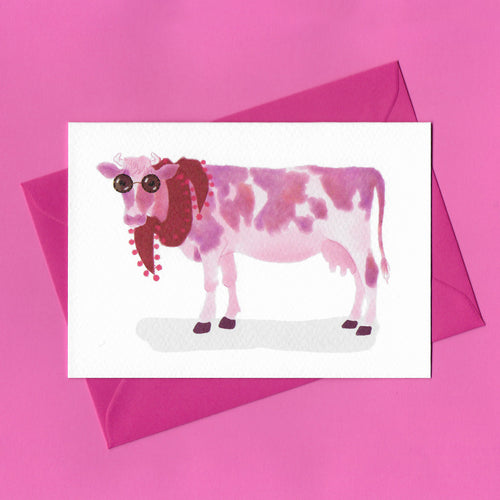 A stylised watercolour illustration on a greeting card of a pink and white cow, wearing sunglasses and a red scarf, with a bright pink envelope on a pink background