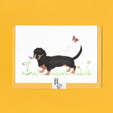 Load image into Gallery viewer, A stylised watercolour illustration on a luxury greeting card of a Black and Tan dachshund dog amongst grass and wildflowers and a butterfly, with a bright yellow envelope held together with an eco friendly plastic free card catch sticker, on a yellow background
