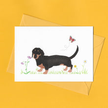 Load image into Gallery viewer, A stylised watercolour illustration on a luxury greeting card of a Black and Tan dachshund dog amongst grass and wildflowers and a butterfly, with a bright yellow envelope, on a yellow background
