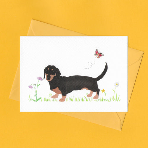 A stylised watercolour illustration on a luxury greeting card of a Black and Tan dachshund dog amongst grass and wildflowers and a butterfly, with a bright yellow envelope, on a yellow background