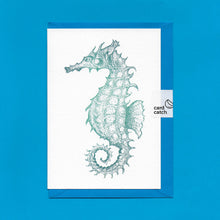 Load image into Gallery viewer, A detailed colour illustration on a luxury greeting card of a teal coloured seahorse, with a kingfisher blue envelope held together with an eco friendly plastic free card catch sticker, sitting on a blue background
